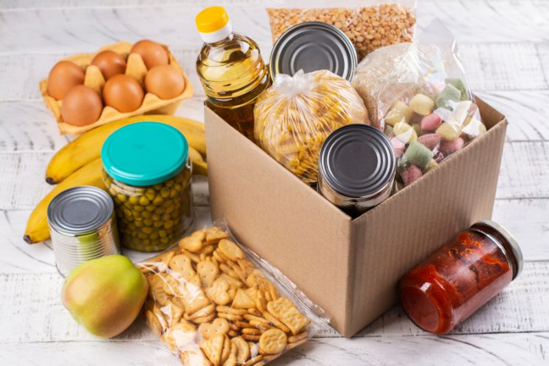 Photograph of eggs and various canned, tinned and other foods in and beside a cardboard box on a white surface.